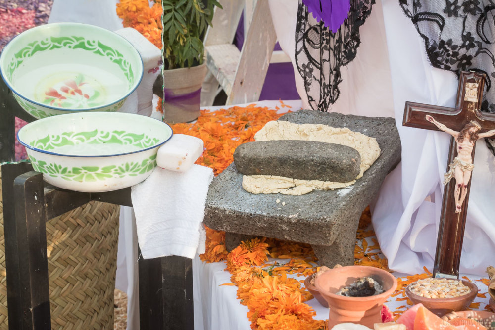 A metate sits next to a cross, a bowl of water with soap and cloth, and a mirror.