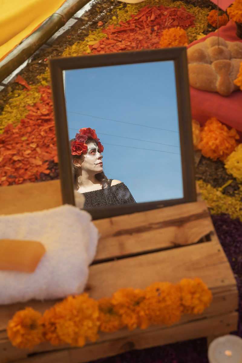 A woman dressed as a catrina is reflected in a mirror on a Day of the Dead altar.