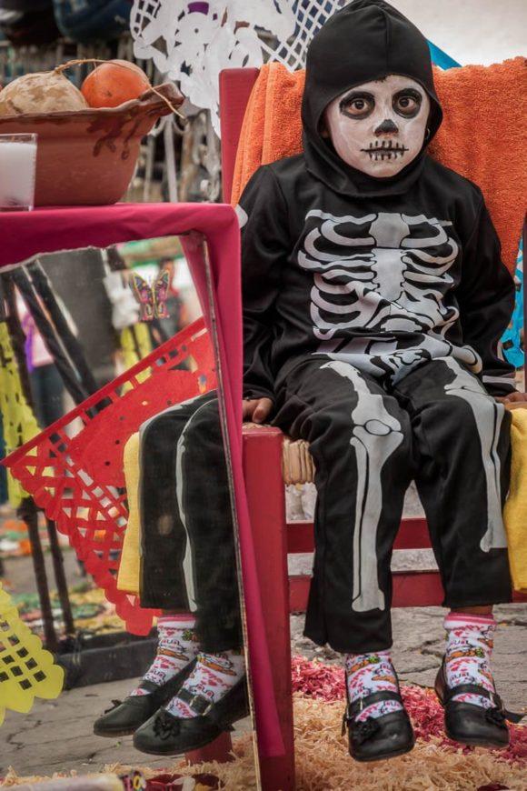 A girl sits as a catrina next to a mirror, a traditional component of the Day of the Dead altar so the spirits can check their appearance upon arrival.