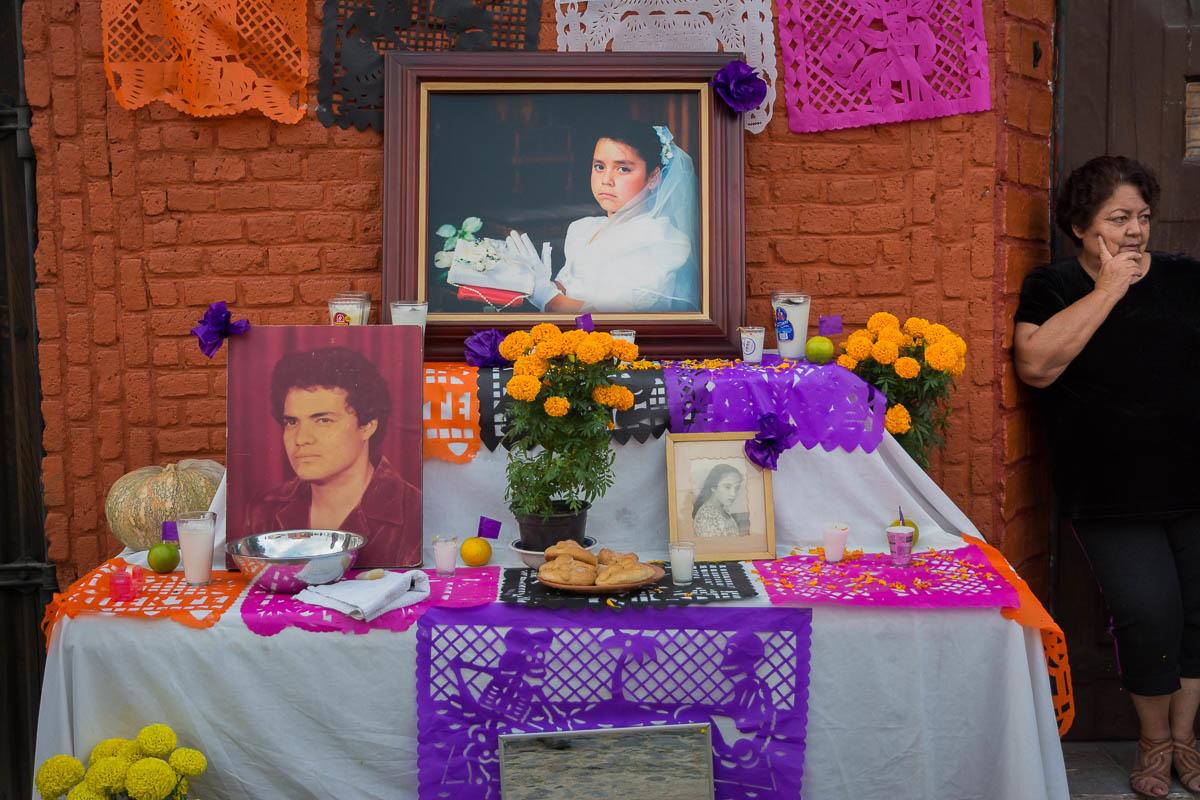 A woman stands beside a Día de los Muertos altar, her familial resemblance to the portraits quite notable. Chapala, Jalisco.